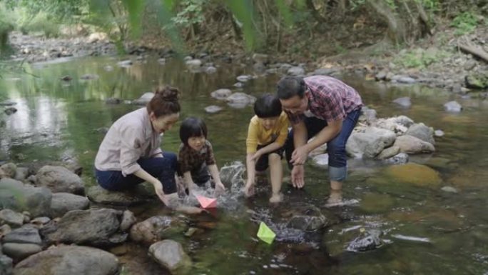 慢动作，一个亚洲家庭，一个父亲和一个母亲教他们的两个儿子的孩子如何制作纸船。漂浮在天然森林溪流中。度