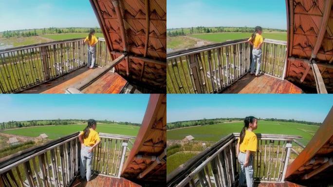 亚洲小女孩站在泰国乡间别墅的阳台上，稻田里有自然的天空。