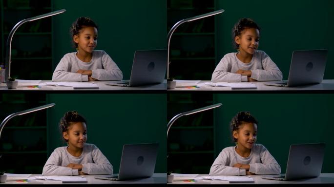 一个女孩在夜灯的灯光下坐在桌子旁，通过笔记本电脑上的视频聊天说话。孩子挥挥手，点了点头，说再见。特写