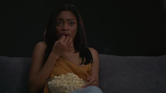 害怕的非洲女人看恐怖电影，晚上在家沙发上吃爆米花。害怕面部表情。家庭影院