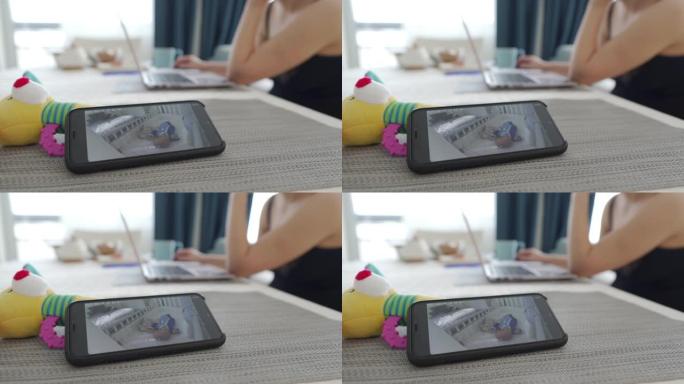 母亲使用手机上的视频婴儿监视器应用程序在笔记本电脑上工作，女人在智能手机屏幕上实时观看睡着的婴儿