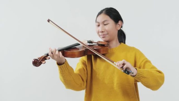 笑着的亚洲女学生穿着黄色毛衣学习拉小提琴与灰色背景，生活理念。