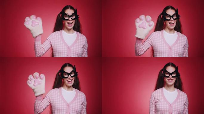 友好的女孩在猫面具有趣而可爱的挥手，并在红色背景的镜头前打招呼。猫女搞笑角色扮演化妆舞会