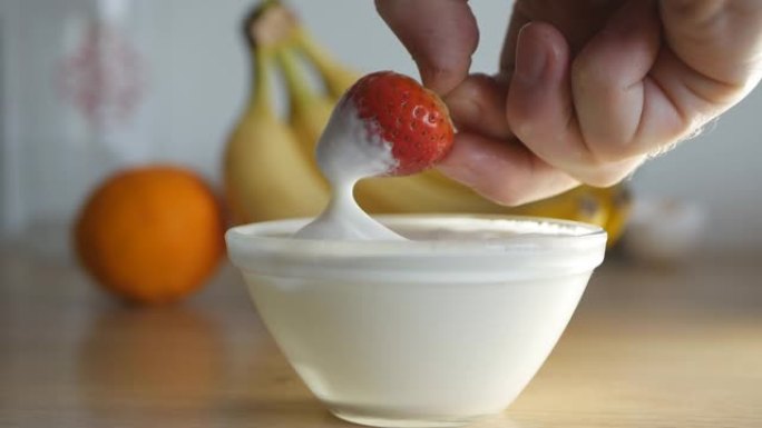 该男子用右手将成熟的红色草莓浸入一碗鲜奶油中，然后食用。背景是水果香蕉和柑橘。关闭4k