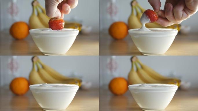 该男子用右手将成熟的红色草莓浸入一碗鲜奶油中，然后食用。背景是水果香蕉和柑橘。关闭4k
