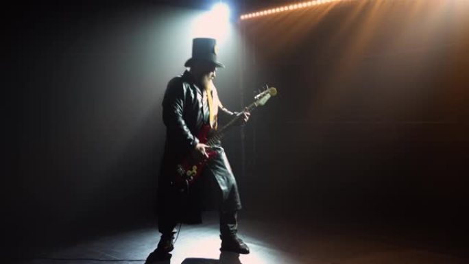 大胡子的摇滚贝斯手在演播室表演中与烟雾和霓虹灯一起独奏。戴着帽子和长长的皮革斗篷的男人的剪影弹吉他。