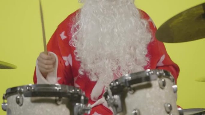 圣诞老人穿着节日服装，戴着胡须和太阳镜，在黄色工作室背景下打鼓。穿着狂欢节服装的人刘海鼓和打击钹来创