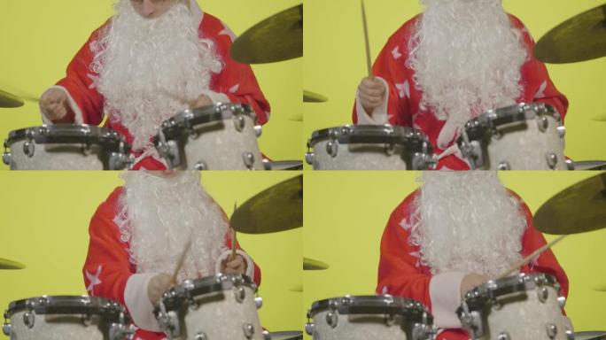圣诞老人穿着节日服装，戴着胡须和太阳镜，在黄色工作室背景下打鼓。穿着狂欢节服装的人刘海鼓和打击钹来创