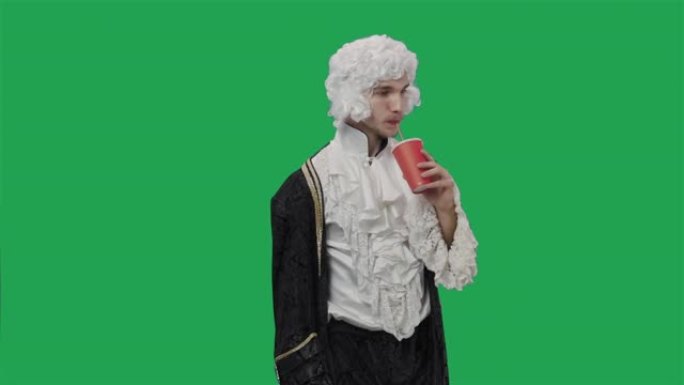 穿着复古西装和假发的朝臣绅士肖像，用塑料玻璃通过吸管喝饮料。年轻人在工作室摆姿势，绿屏背景。特写。慢