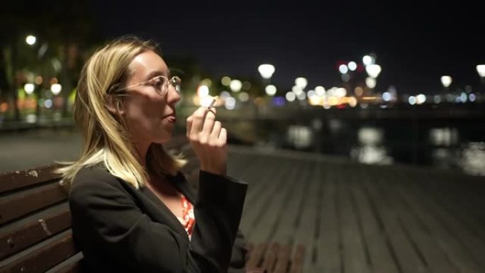 经过漫长而劳累的一天，疲惫的现代年轻女子坐在码头的长凳上，晚上抽烟