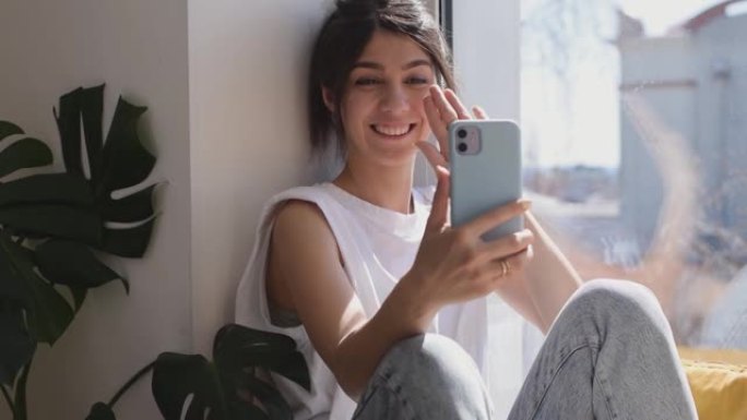 一个黑发混血女孩正坐在窗台上的家里，在智能手机上的视频链接上进行情感交谈。现代技术。