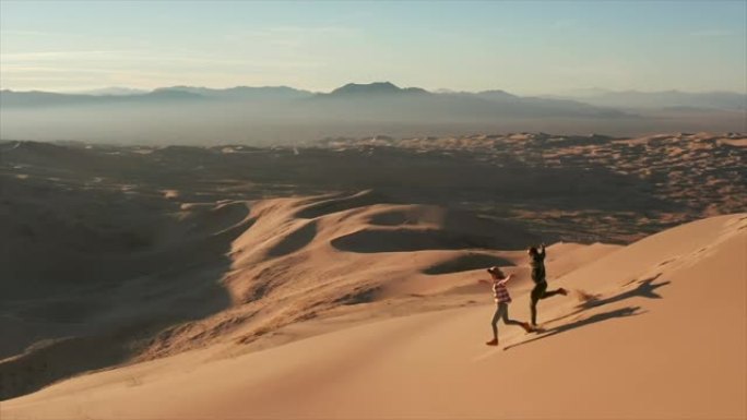 跟踪镜头，一对夫妇在日落时跑下理智的沙丘。慢动作的人在沙漠中的圣沙丘上奔跑