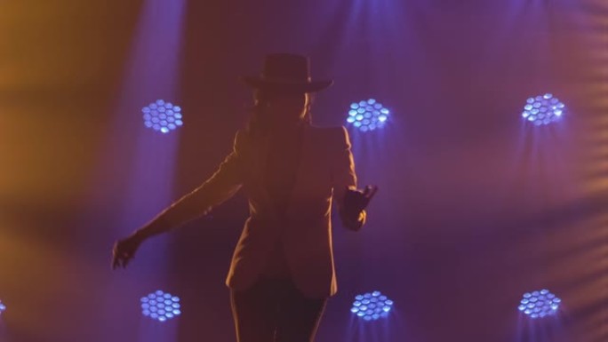 穿着长裤套装和帽子的迷人女人的剪影，在蓝色工作室灯光下的黑暗中表演阿根廷弗拉门戈舞蹈编舞。西班牙舞者