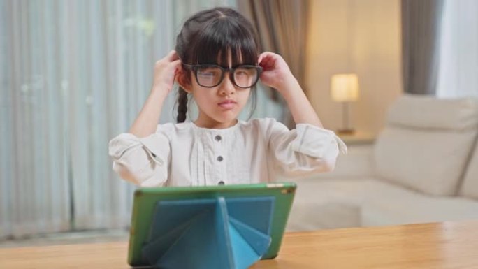 亚洲迷人的小可爱孩子坐在桌子上，看卡通或在家里的客厅平板电脑上学习。年轻的女学生戴眼镜保护视力从数字
