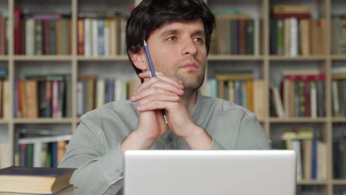 体贴的年轻人坐在办公室的办公桌前，手里拿着一支铅笔，移开目光，在笔记本电脑上休息，思考项目计划