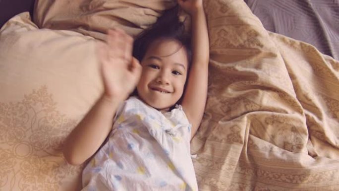穿着睡衣的白色皮肤可爱的亚洲小孩的肖像，早上在床上对着镜头，阳光透过窗户照射。她的眼神交流表达了喜悦