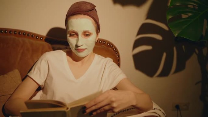 美国妇女正在沙发上读书，并在面部spbas上戴着化妆品面膜