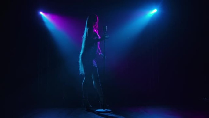 流行歌手在蓝色紫罗兰色灯光照亮的黑暗舞台上表演的仰视图。在现场音乐会上，一头长发飘逸的女人的深色剪影