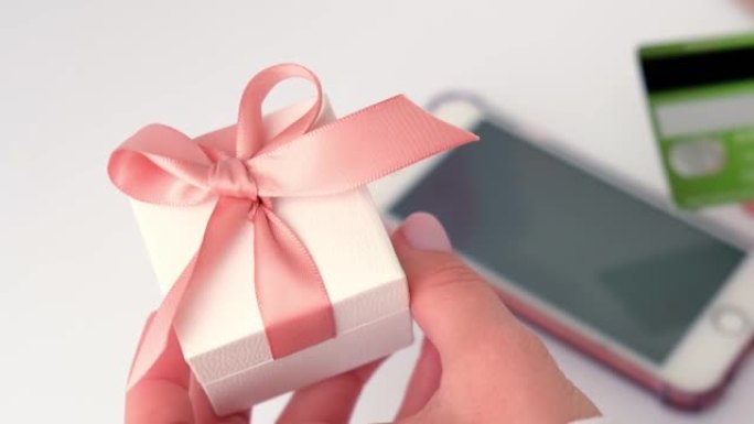 使用智能手机上的在线银行应用程序并在家中持有借记卡的女性手持礼品礼物盒的特写视图。网上购物。假期礼物