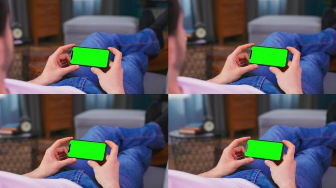 在家中的年轻人在水平景观模式下使用绿色模拟屏幕手机在沙发上休息。男孩正在浏览互联网，观看视频，社交媒