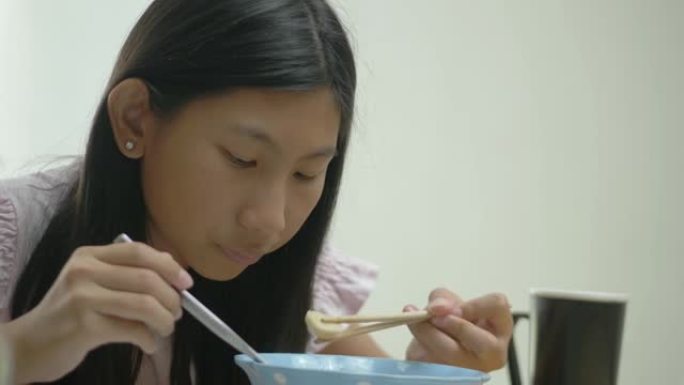 亚洲女孩吃面条汤，在家里的餐桌上通过智能手机看电影或病毒视频，生活方式概念。
