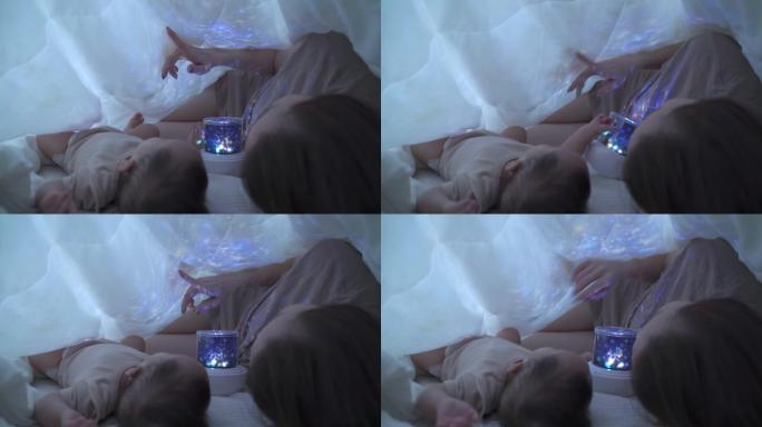 亚洲女性母亲父母在家卧室用毯子下的婴儿玩星空夜光投影仪。点星睡前教宝宝一堂课。