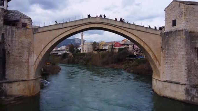 人们在古镇两岸之间的山河上的历史拱形石桥上行走，在桥跨下多云的天空飞行下