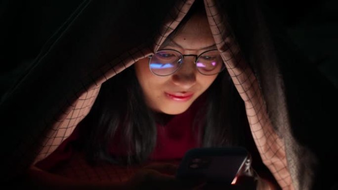 亚洲女性在家里卧室的黑暗中使用羽绒被下的智能手机感到高兴。
