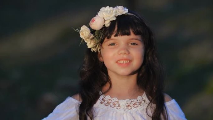 一个白人学龄前小女孩的特写肖像，棕色长发，蓝色的眼睛在花环上跳舞，在自然背景上微笑。4k 50 fp