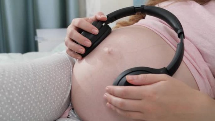 在孕妇的大肚子上播放音乐的大耳机的平移镜头。未出生的婴儿在母亲腹部听经典音乐。早期艺术和创造力发展的