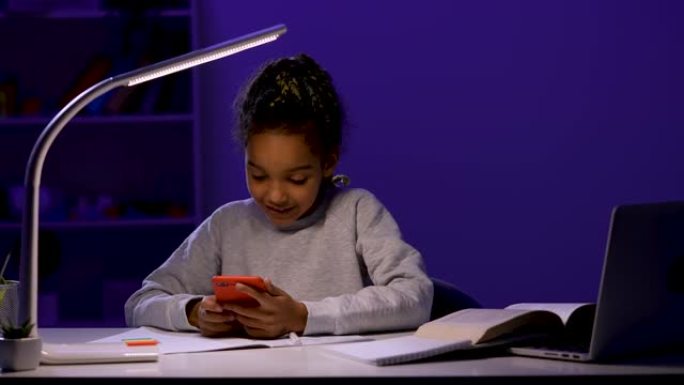 一名女学生写作业，拿起手机检查信息并回复，然后继续学习。在夜灯的灯光下坐在桌子上的孩子。特写。慢动作