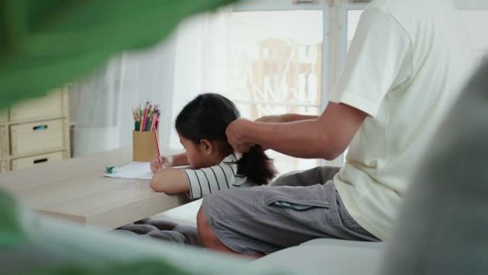 亚洲女孩在做作业，父亲在客厅里梳头编织头发。