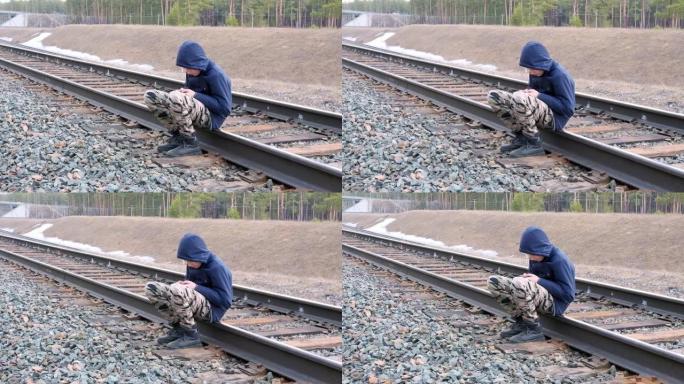 孤独的少年坐在危险中的铁轨上。男孩歪着头玩电话，没有注意到周围的任何东西