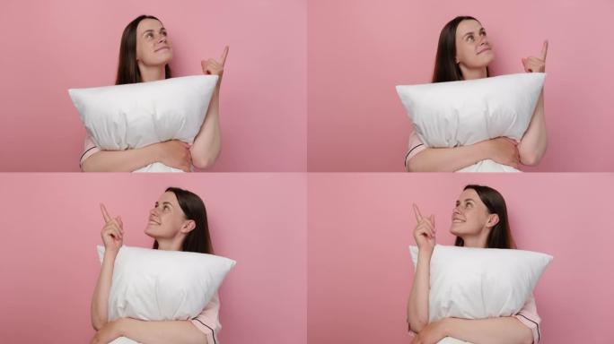 穿着睡衣的欢快的梦dream以求的年轻女子在家休息时，用食指举起白色枕头，在柔和的粉红色工作室背景墙