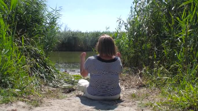 穿着便服和帽子的活跃快乐健康的欧洲老年妇女坐在湖边的莲花位置，举起手来冥想。瑜伽，安心，精神卫生概念