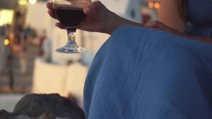 坐在Oia城堡上的小玻璃杯中拿着自制的甜玫瑰酒的女人的局部视图和坐在Oia城堡墙上的希腊岛圣托里尼岛