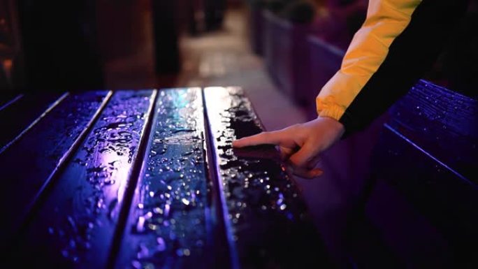 那家伙沿着街道上潮湿的桌子跑手指，从桌子上搬走水。慢动作拍摄特写。周围都是霓虹灯