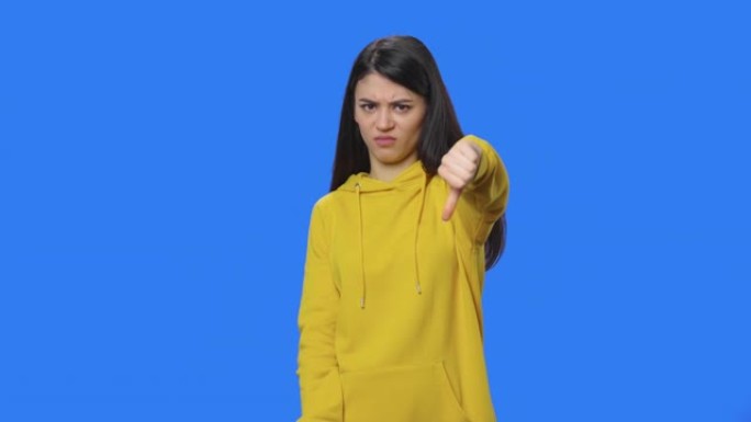 漂亮黑发女人的肖像显示拇指朝下的手势。穿着黄色运动衫的长发年轻女子在蓝屏背景的工作室摆姿势。特写。慢