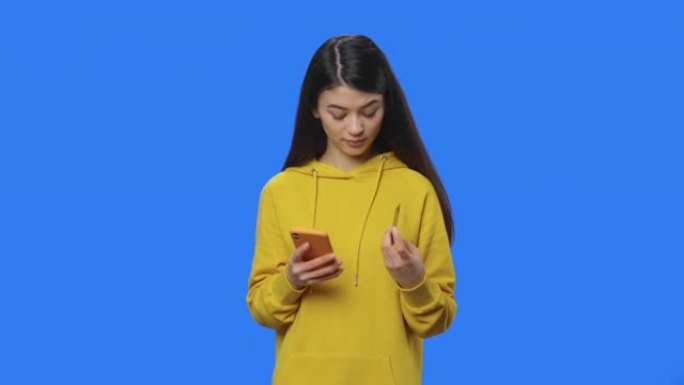 漂亮黑发肖像使用信用卡和电话在线购买。穿着黄色运动衫的年轻女子在蓝屏背景的工作室摆姿势。特写。慢动作