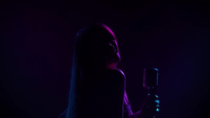 迷人的女歌手在派对上用复古麦克风表演现场音乐。在黑暗的工作室里，一位长发被蓝色和紫色灯光照亮的艺术家