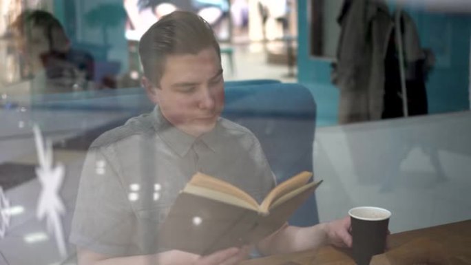 一个年轻人坐在咖啡馆的玻璃杯后面看书。这个人正在喝咖啡。