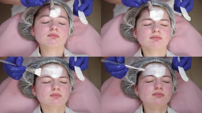 美容师用化妆刷在客户脸上涂抹白色清洁脱皮泥面膜。皮肤科诊所接受皮肤治疗的年轻女孩的特写