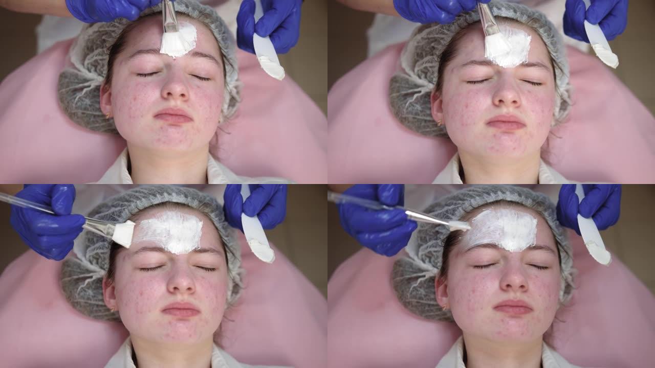美容师用化妆刷在客户脸上涂抹白色清洁脱皮泥面膜。皮肤科诊所接受皮肤治疗的年轻女孩的特写
