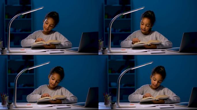一名非裔美国女学生在夜灯的灯光下做作业，大声朗读一本书。孩子坐在家里的笔记本电脑前的桌子旁。特写。慢