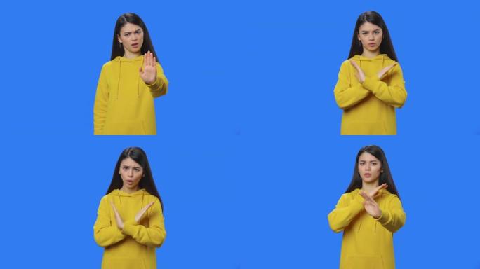 布鲁内特的肖像用手严格打手势，这意味着否认说不。穿着黄色运动衫的年轻女子在蓝屏背景的工作室摆姿势。特