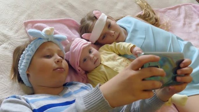 孩子们躺在床上，带着婴儿在智能手机上观看有趣的视频