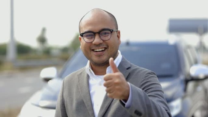年轻的亚洲企业穿着西装站着，在车前竖起大拇指。年轻人得到新车他站在路边。