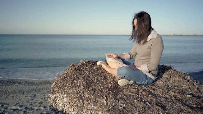 年轻的黑发女人坐在荒凉的海滩上，享受着淡季海洋的宁静，读一本纸质书。体验自然远离现代的狂热和技术习惯