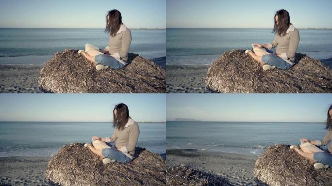 年轻的黑发女人坐在荒凉的海滩上，享受着淡季海洋的宁静，读一本纸质书。体验自然远离现代的狂热和技术习惯
