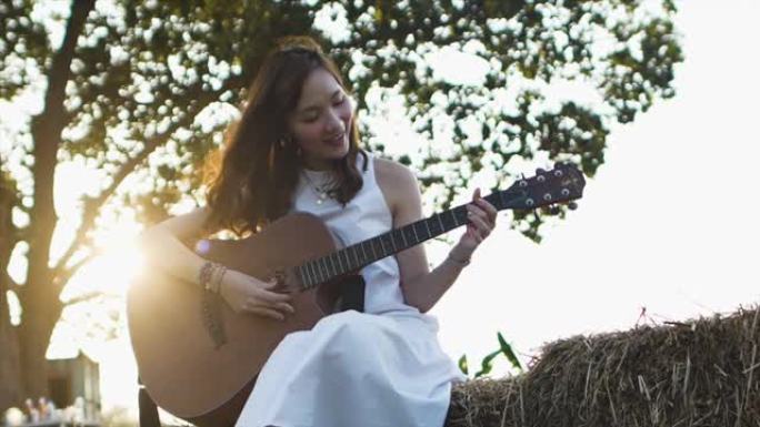 慢动作一位穿着白色连衣裙的年轻年轻亚洲女子在清晨的阳光镜头耀斑下在干草堆上弹吉他。